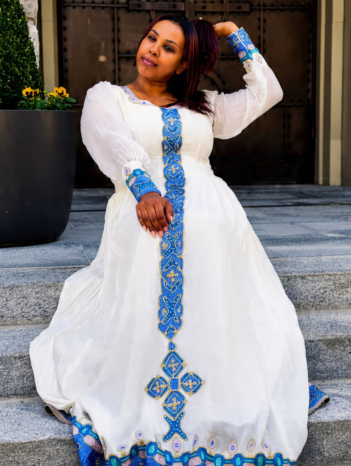 A woman wearing a white traditional Habesha Store  with intricate blue and gold embroidery, posing on stone steps in front of a building entrance.