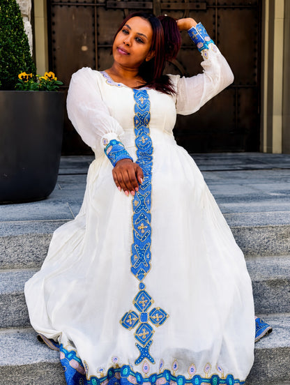 A woman wearing a white traditional Habesha Store  with intricate blue and gold embroidery, posing on stone steps in front of a building entrance.