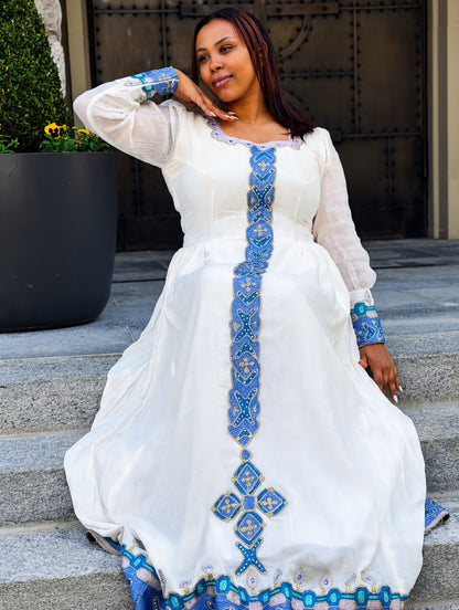 A woman wearing a white long-sleeve Habesha Store  with ornate blue and gold embroidery, sitting on stone steps in front of a dark metal gate.