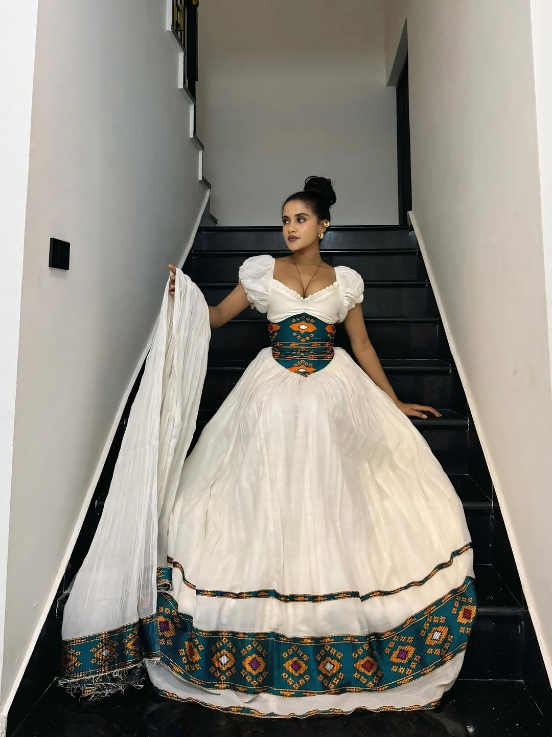 Woman in a white Ethiopian cultural dress with intricate patterns standing on a staircase.