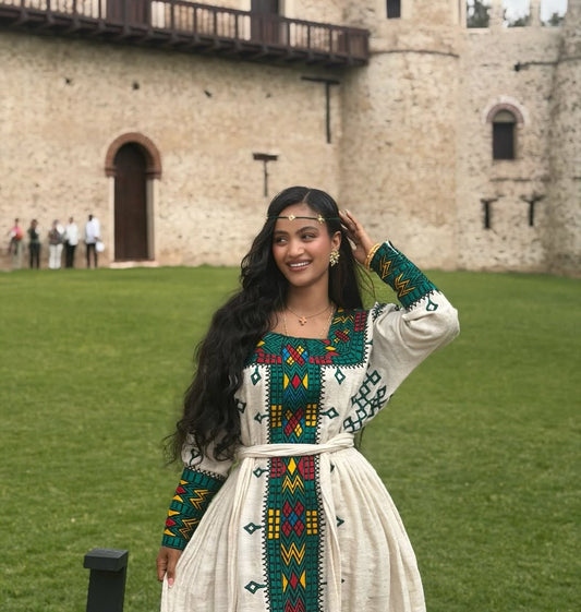 Woman in Ethiopian Traditional Dressstanding in front of a stone building with green grass.