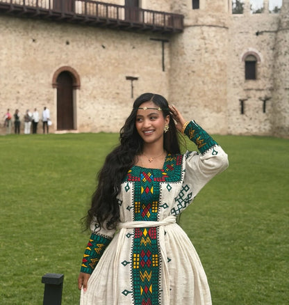 Woman in Ethiopian Traditional Dressstanding in front of a stone building with green grass.