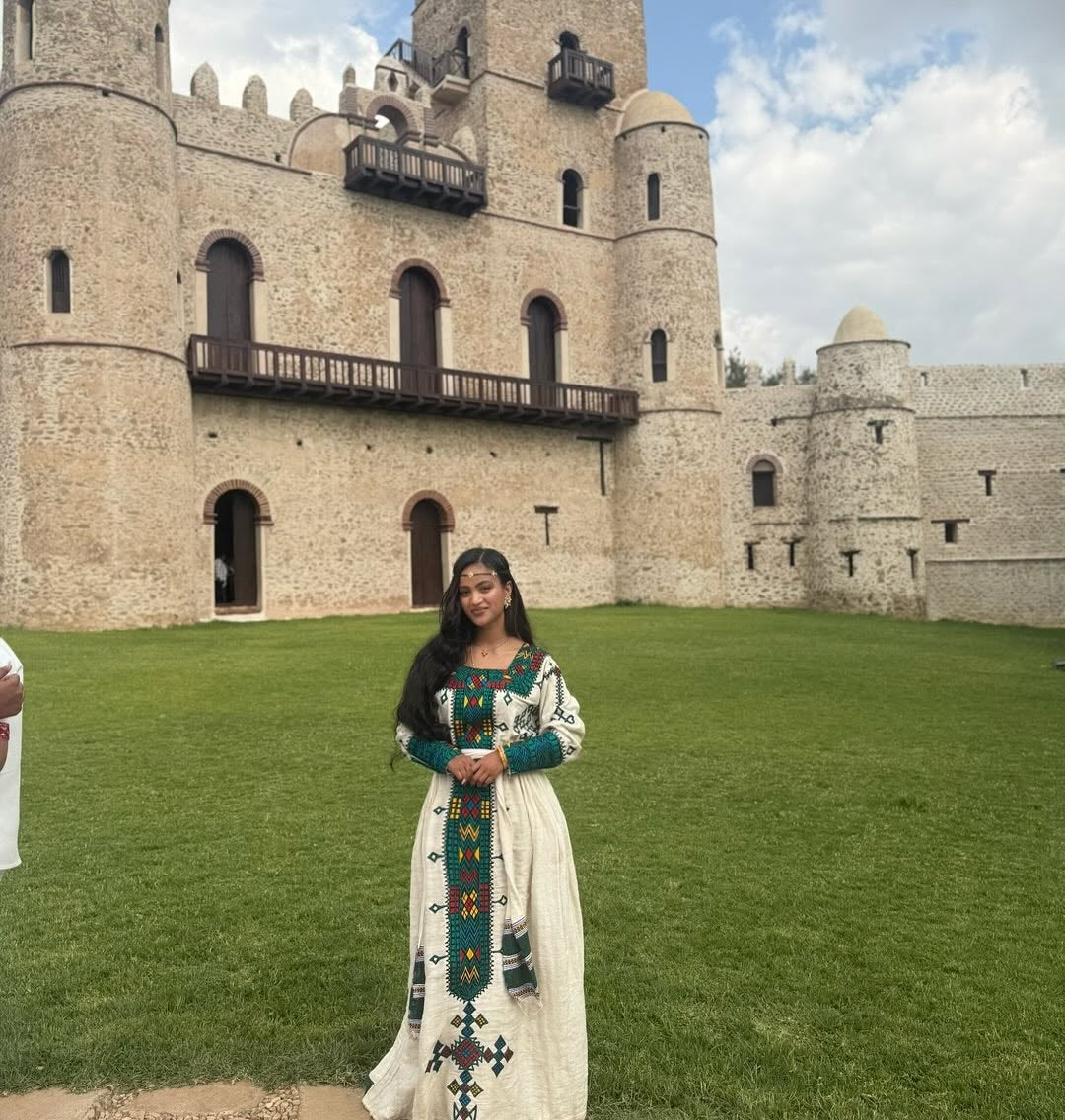 Woman in Ethiopian Traditional Dress attire standing in front of a large stone building with a green lawn.