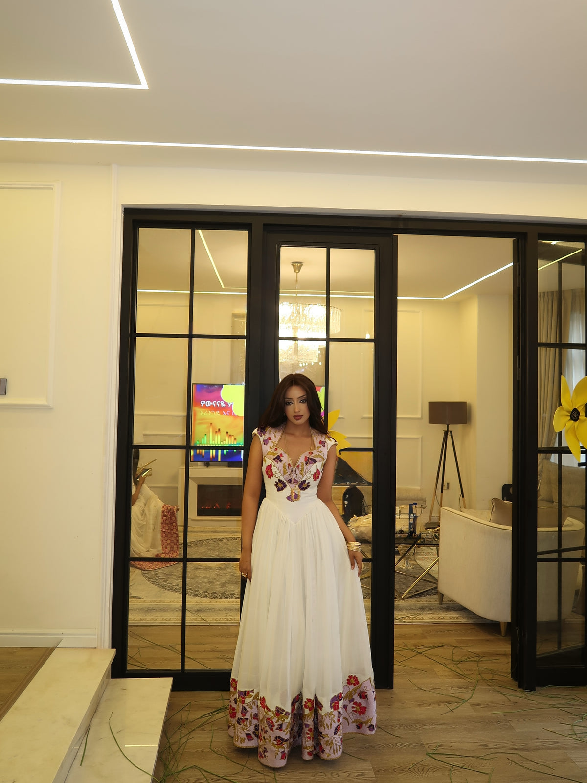 A woman wearing a white sleeveless maxi Habesha store dress with colorful floral embroidery on the bodice and hem, standing in front of black-framed glass doors in a modern indoor setting.