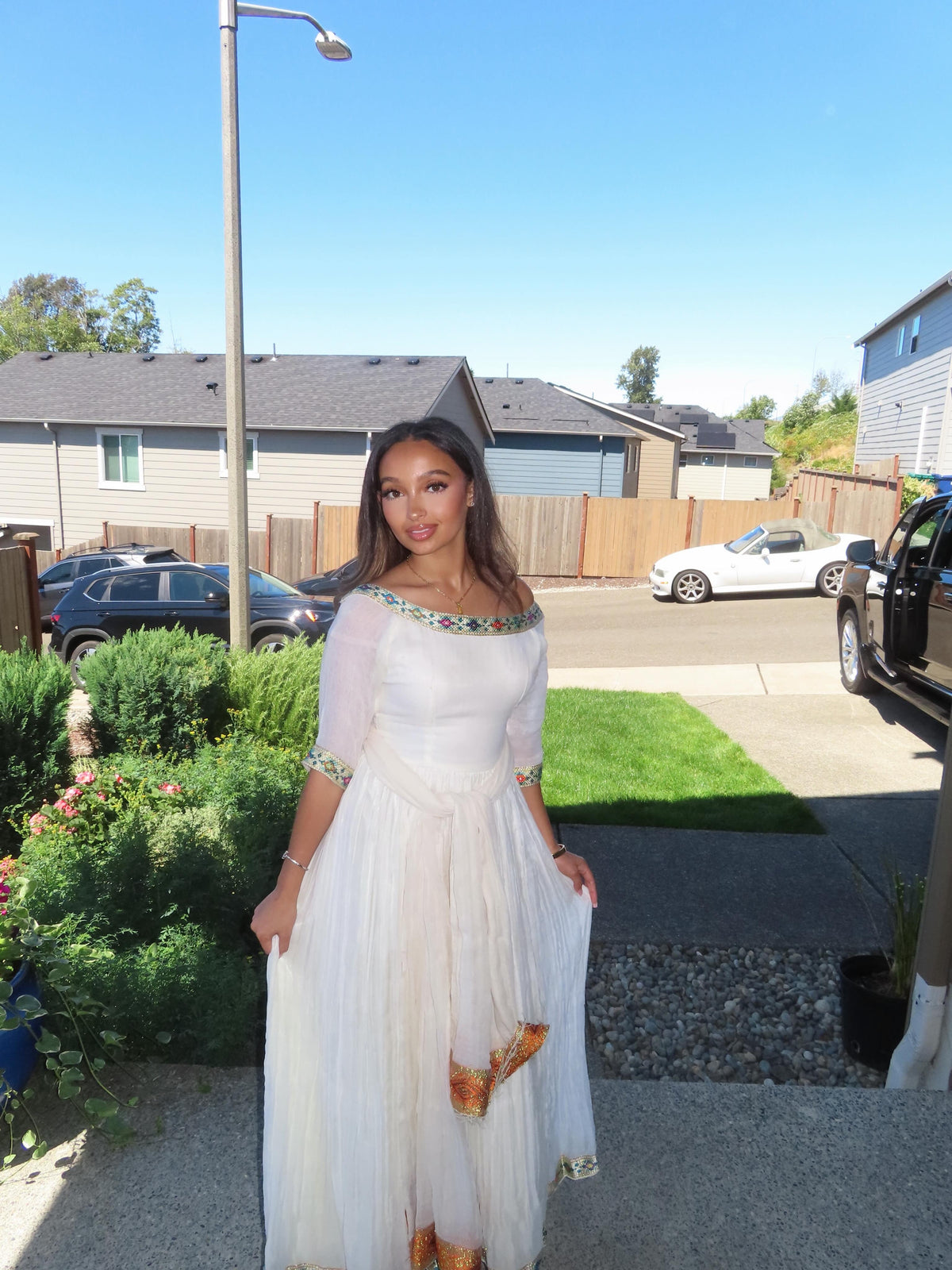 A woman wearing a white off-the-shoulder maxi Ethiopian Store Dress with gold and green beaded trim and gold patterned accents near the hem, standing outdoors in a residential neighborhood.
