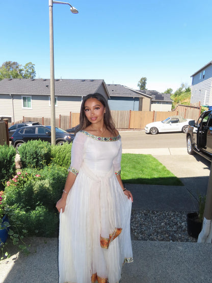 A woman wearing a white off-the-shoulder maxi Ethiopian Store Dress with gold and green beaded trim and gold patterned accents near the hem, standing outdoors in a residential neighborhood.