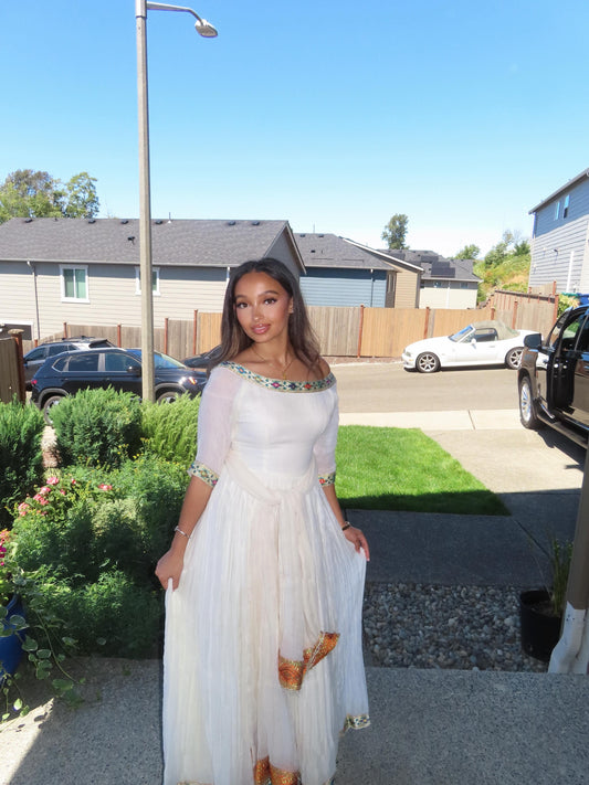 A woman wearing a white off-the-shoulder maxi Ethiopian Store Dress with gold and green beaded trim and gold patterned accents near the hem, standing outdoors in a residential neighborhood.
