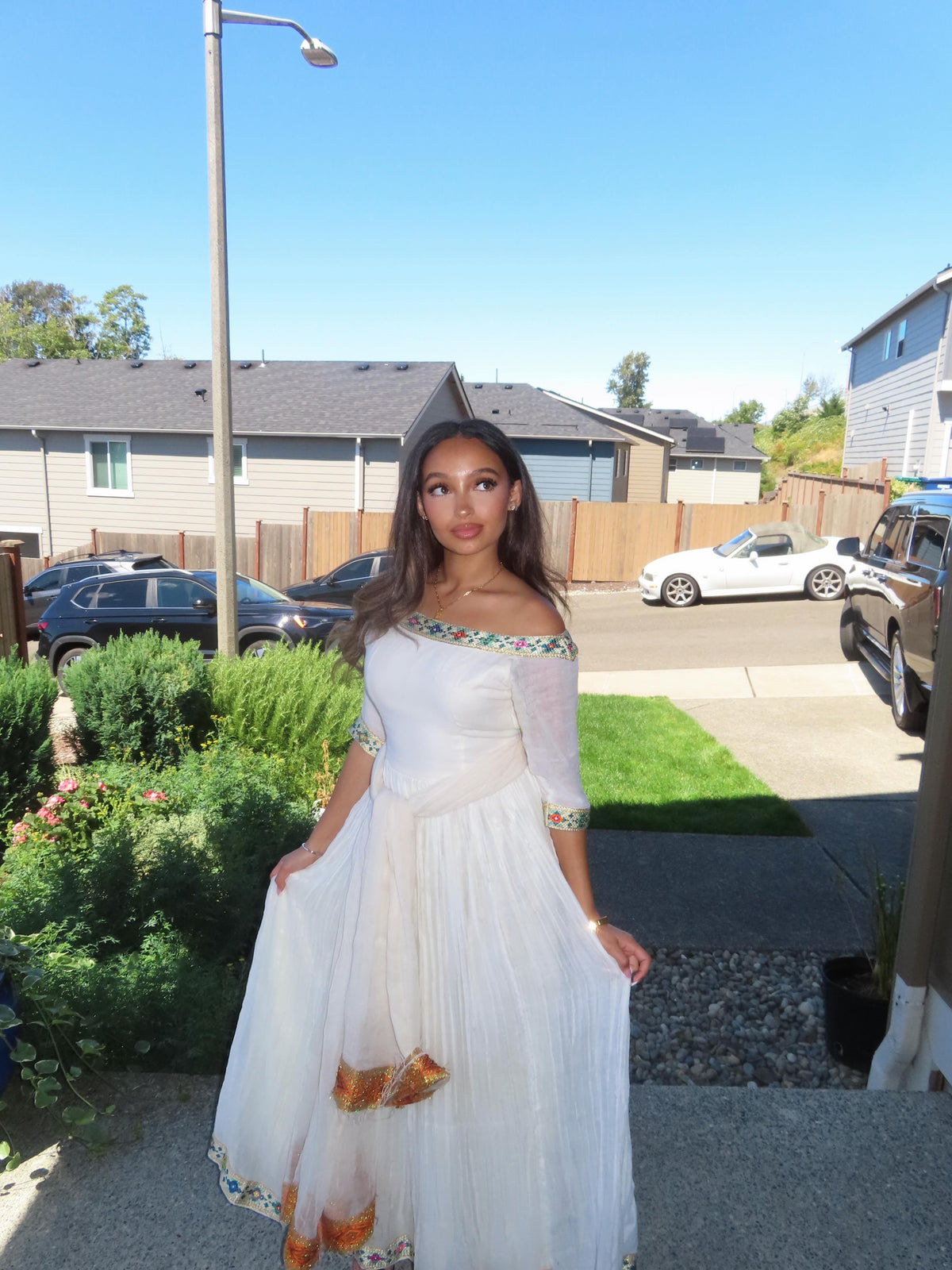 A woman wearing a white off-the-shoulder maxi Ethiopian Store Dress with colorful embroidered details and a gold pattern near the hem, standing outdoors in a residential neighborhood.
