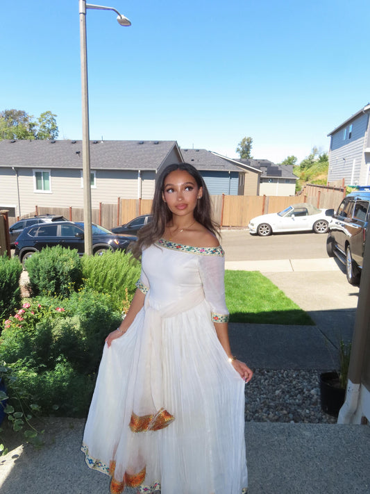 A woman wearing a white off-the-shoulder maxi Ethiopian Store Dress with colorful embroidered details and a gold pattern near the hem, standing outdoors in a residential neighborhood.