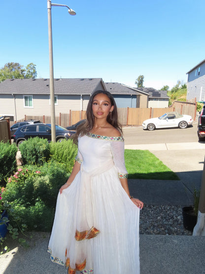 A woman wearing a white off-the-shoulder maxi Ethiopian Store Dress with colorful embroidered details on the bodice and hem, standing outdoors in front of a suburban house.