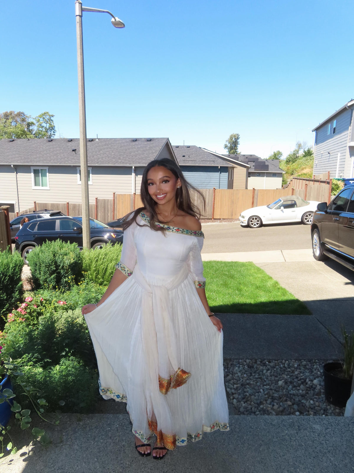 A woman wearing a white off-the-shoulder maxi Ethiopian Store Dress with gold and green embroidered details on the sleeves and hem, standing outdoors in a residential neighborhood.