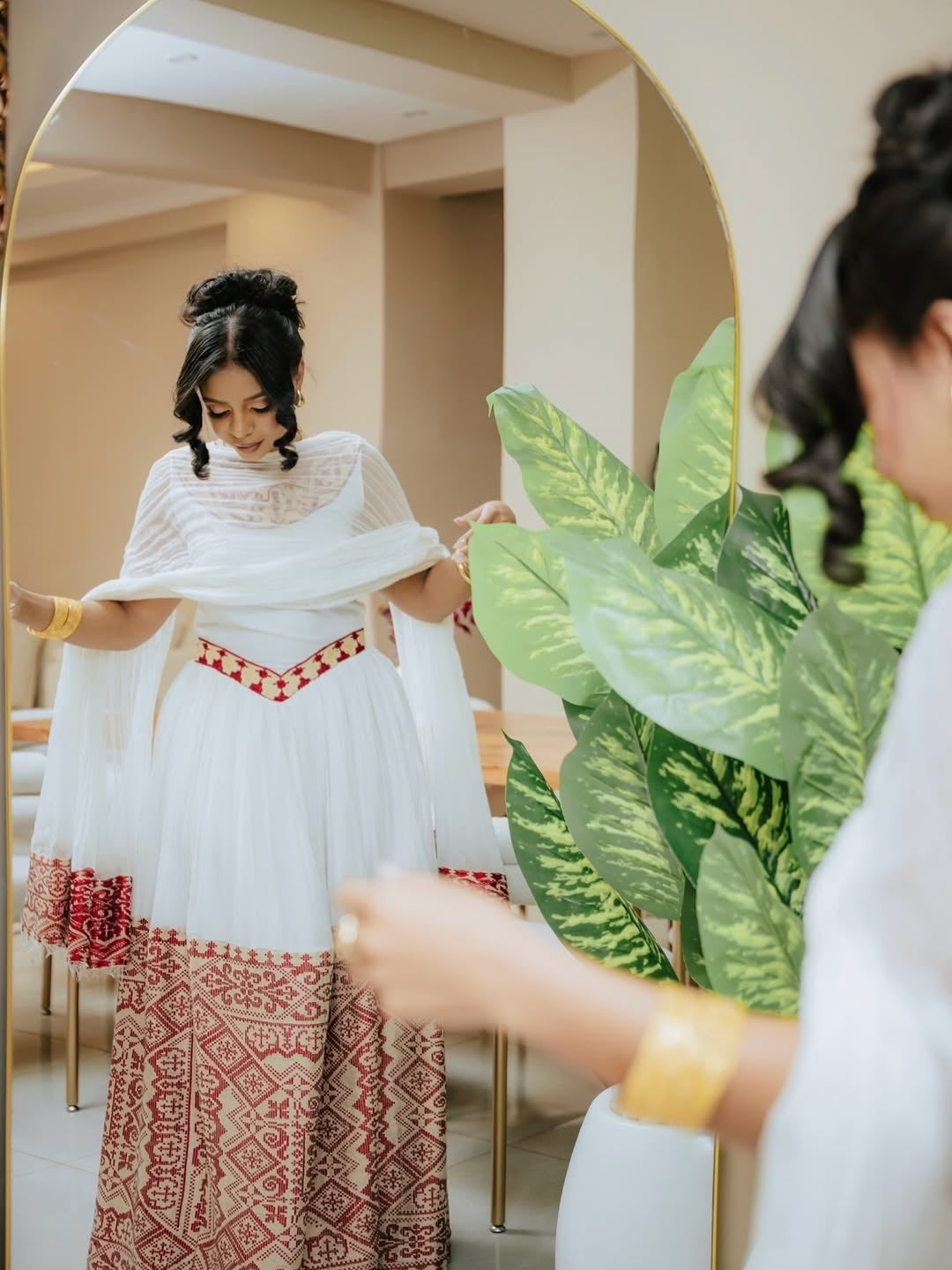 Woman adjusting a white garment with red red Habesha Libs in a room with plants.