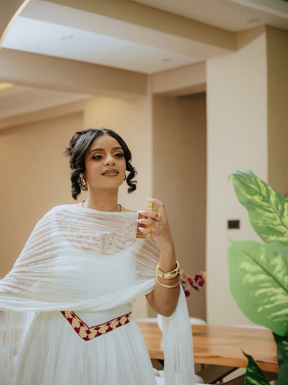 Woman in a white traditional red Habesha Libs holding a glass of water indoors.