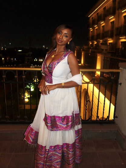 Woman in a colorful habesha dress standing on a balcony at night.