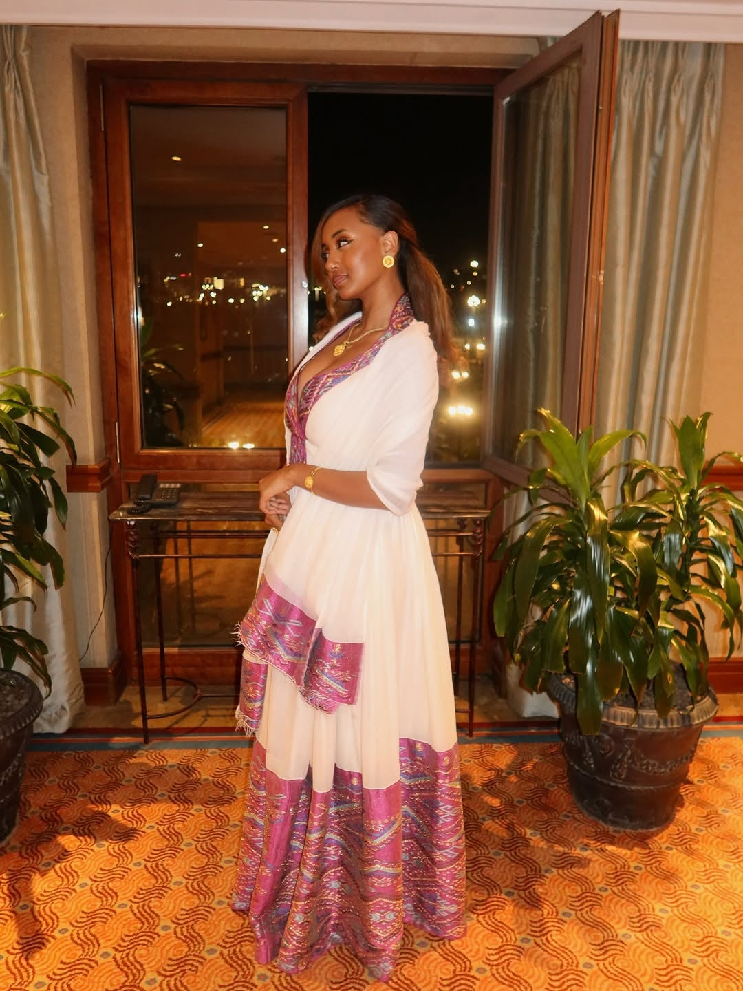 Woman in a white and pink traditional habesha dress standing in a room with plants and a window.