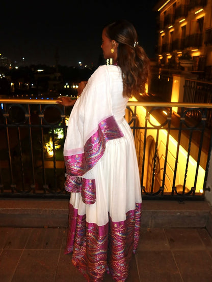 Woman in a white habesha dress with purple and pink patterns standing on a balcony at night.