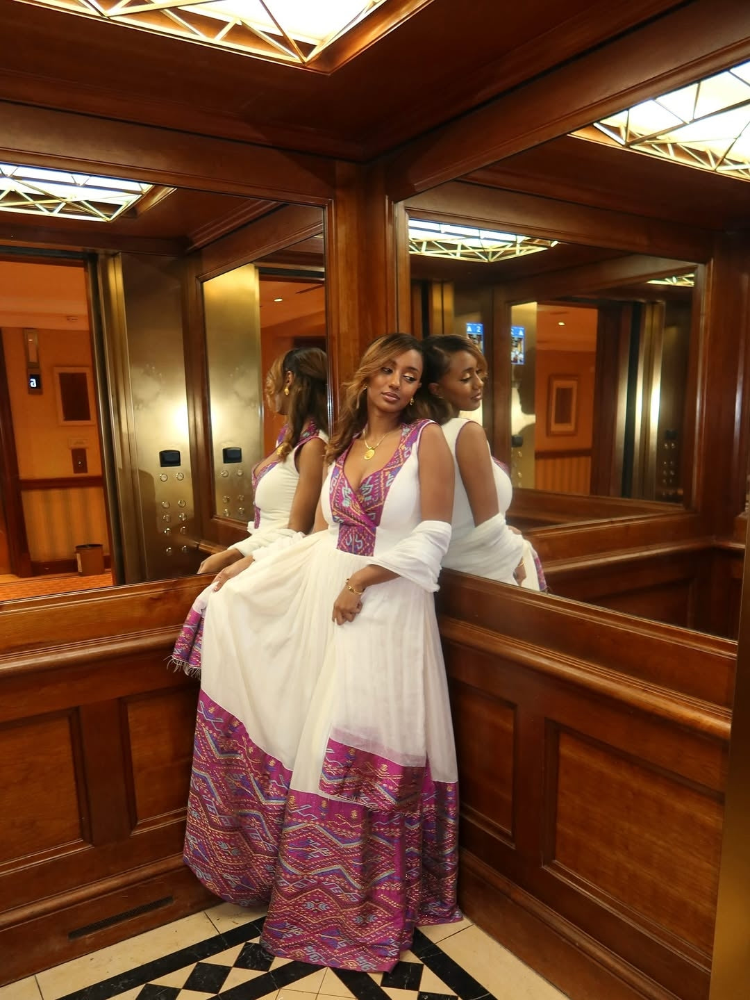 Woman in a white and pink habesha dress standing in a wooden-paneled room with mirrors.