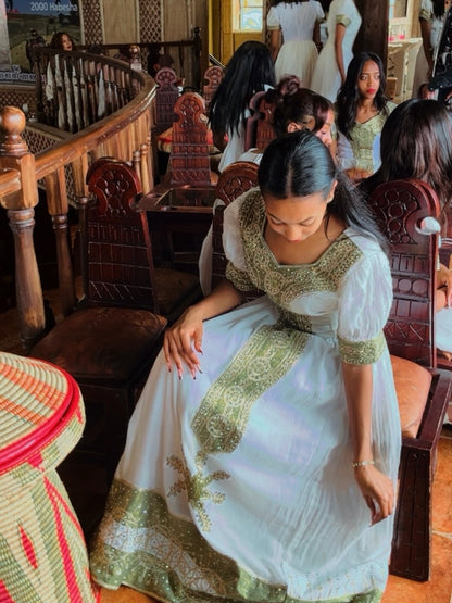 Woman in a white and green dress sitting in a room with wooden chairs and tables.