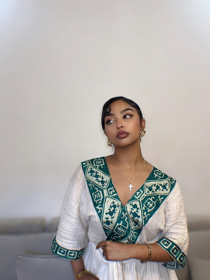 Woman wearing a white and green Ethiopian cultural dress with top against a plain background