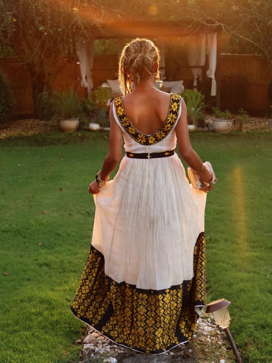 A woman viewed from behind wearing a white Ethiopian dress  with a black and gold geometric patterned border, standing in a grassy backyard at sunset.