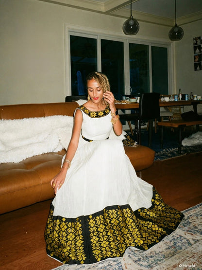 A woman wearing a white and black patterned Ethiopian dress  sits on a brown leather couch in a living room setting.