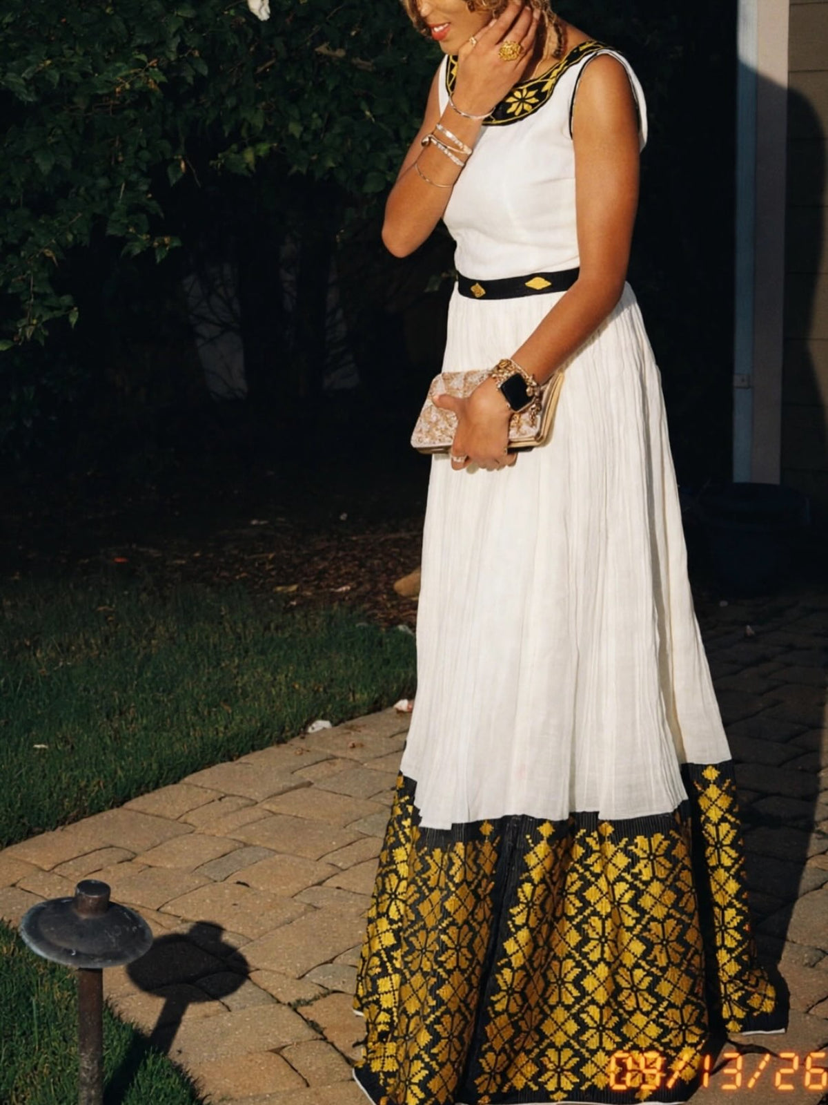 A woman wearing a sleeveless white Ethiopian dress with a black and gold patterned hem and waist detail, standing outdoors on a paved walkway at night.