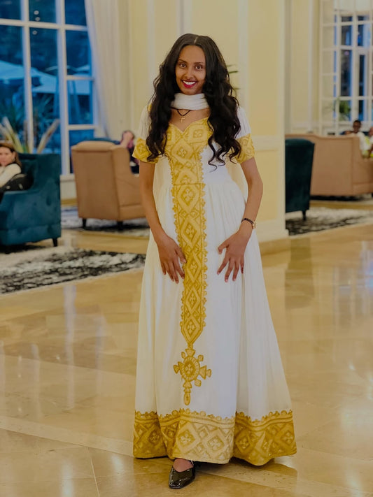 Woman in a white and yellow Habesha kemis standing in a well-lit room with large windows.