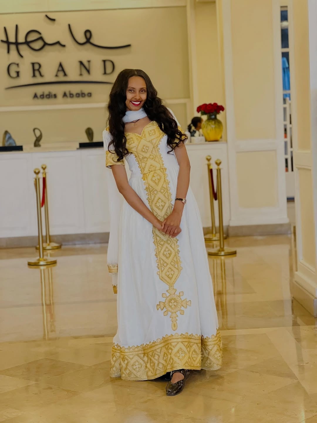 Woman in a white and gold Habesha kemis standing in front of a 'Hailu Grand' sign.