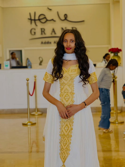 Woman in a white and yellow Habesha kemis standing in a lobby with 'Hätel Grand Addis Ababa' in the background.