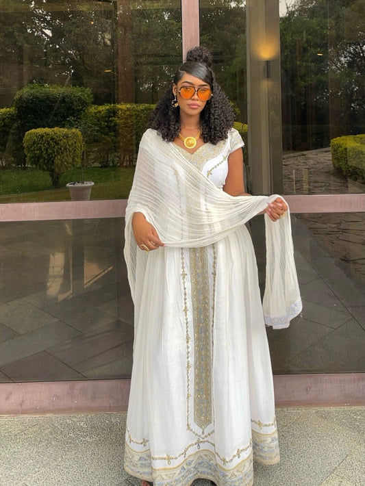 A woman stands outdoors wearing a long white traditional Habesha Store with gold embroidery and a matching draped scarf. She is wearing orange-tinted sunglasses, a gold necklace, and earrings, with a glass patio door and green garden visible in the background.
