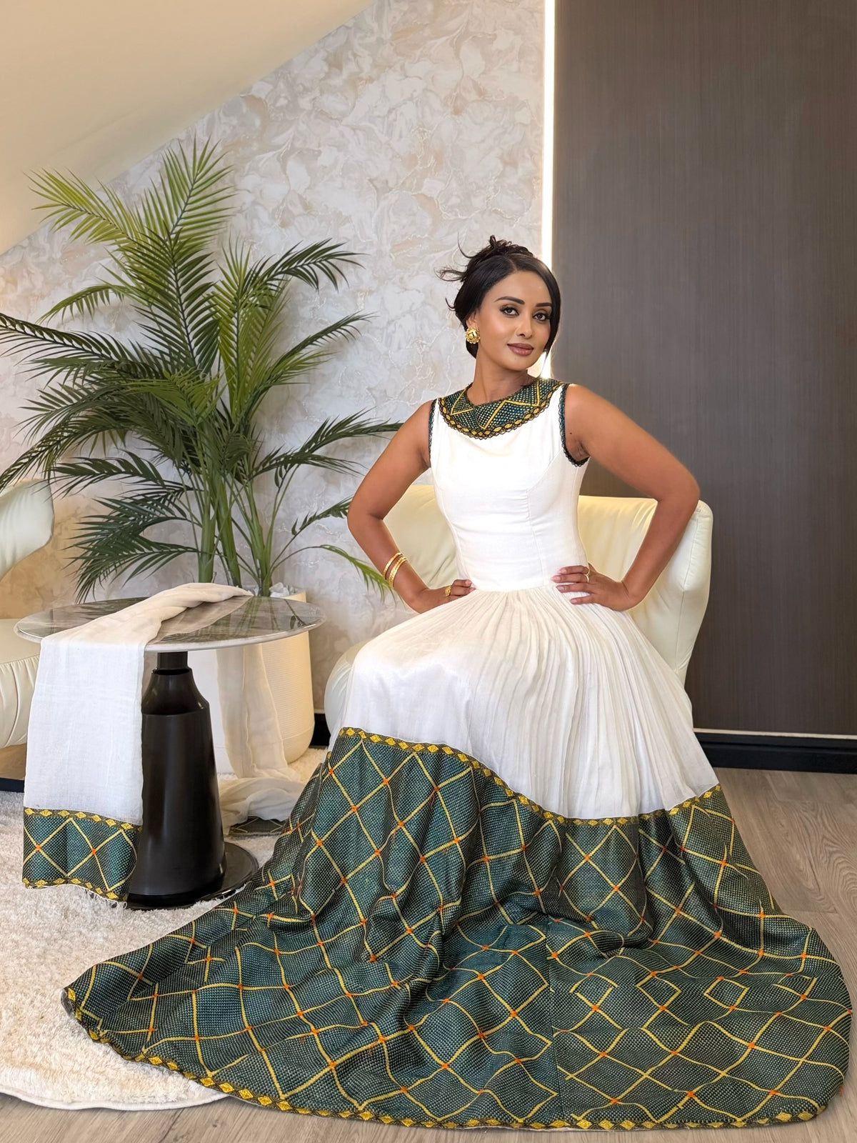 A woman wearing a sleeveless white  Ethiopian dress  with a green and gold patterned skirt and neckline, seated on a white chair in an indoor setting with a plant and side table.