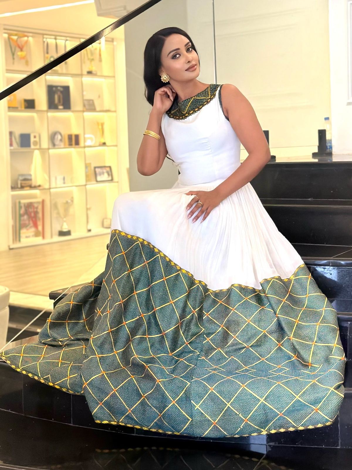 A woman models a sleeveless white and green traditional  Ethiopian dress  with gold geometric patterns and a green embroidered neckline, sitting on a black staircase in an indoor setting.
