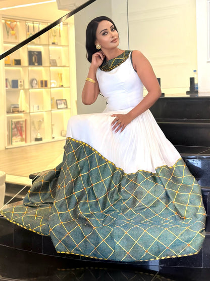 A woman models a sleeveless white and green traditional  Ethiopian dress  with gold geometric patterns and a green embroidered neckline, sitting on a black staircase in an indoor setting.