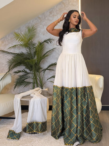 A woman models a white sleeveless maxi  Ethiopian dress  with a green and gold geometric patterned hem and neckline, styled in a modern interior setting with a matching dupatta draped on a side table.