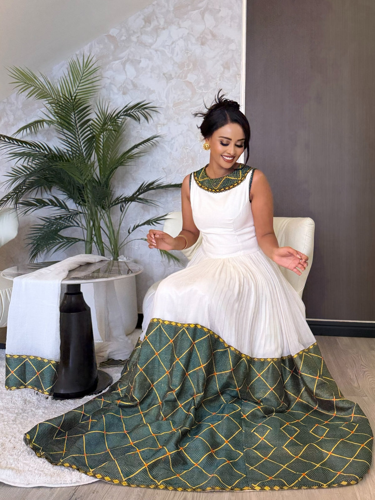 A woman wearing a sleeveless white  Ethiopian dress  with a green and gold geometric patterned skirt and collar, sitting in a white chair in an indoor setting with a potted plant.