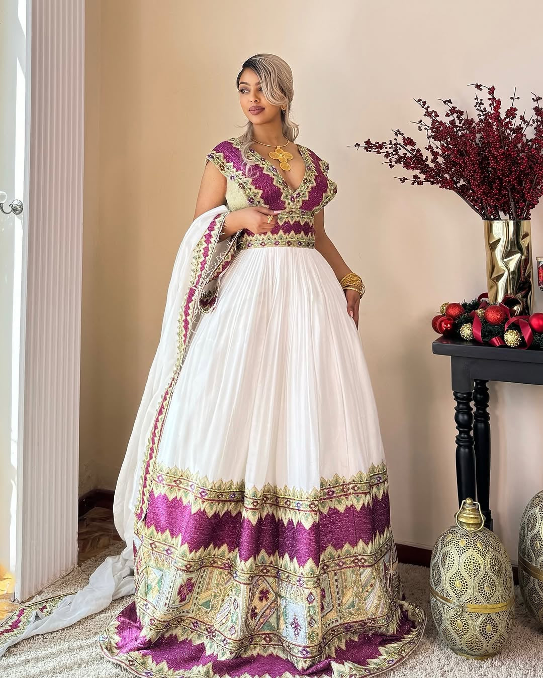 Woman in a traditional Habasha dress   with white and purple patterns, standing indoors.