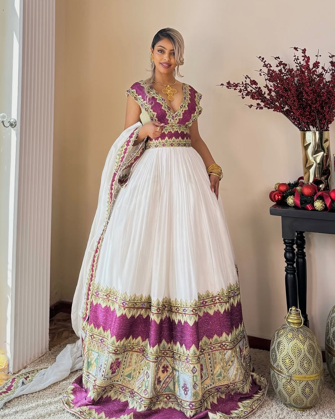Woman in a white and purple traditional habesha kemis  standing indoors with decorative elements.