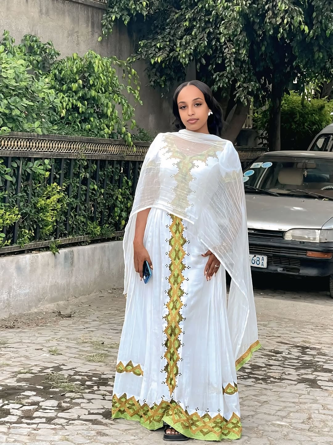 Woman in a white and green traditional Gold Habesha Kemis standing outdoors with greenery in the background.