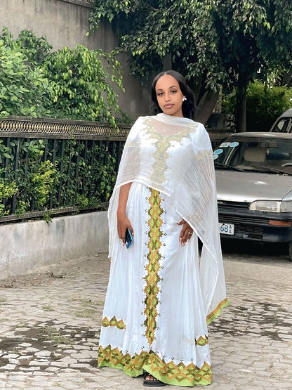 Woman in a white and green traditional Gold Habesha Kemis standing outdoors with greenery in the background.