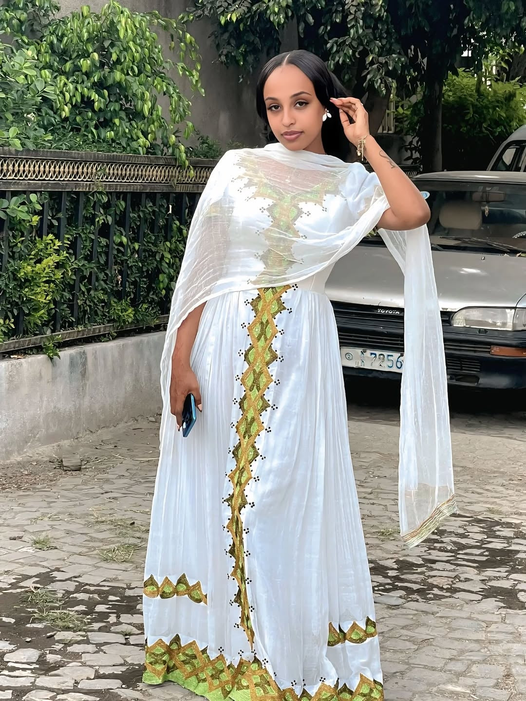 Woman wearing a white Gold Habesha Kemis with green patterns, standing outdoors.