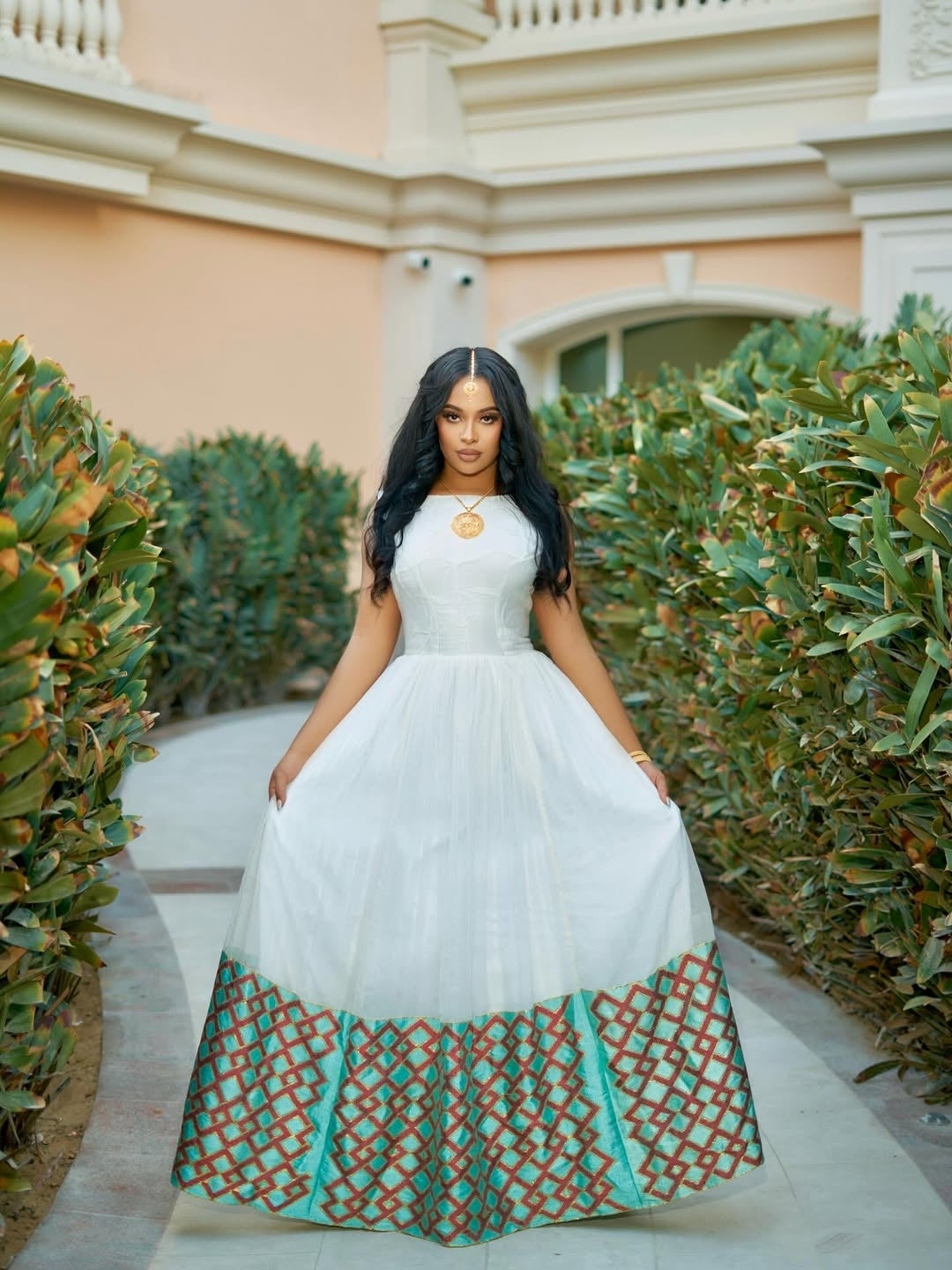 Woman in a white Ethiopian cultural dress  with patterned bottom standing in front of green bushes and a building.