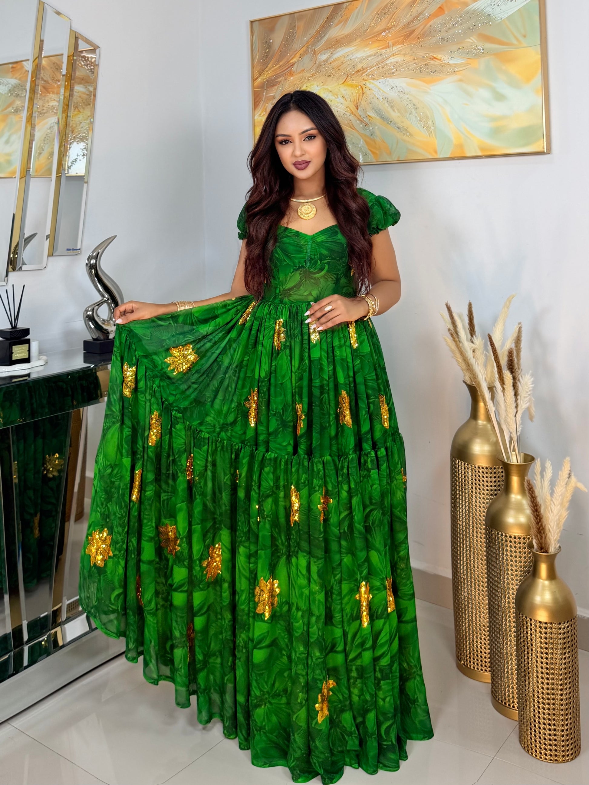 A woman models a vibrant green tiered Habesha Chiffon with gold floral embroidery, standing in an indoor setting with decorative gold vases and a mirror.