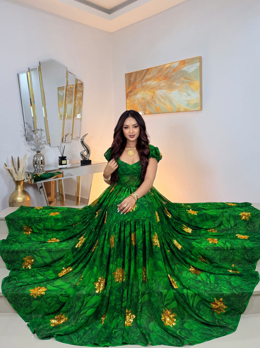 A woman wearing a voluminous emerald green ball Habesha Chiffon with gold floral embroidery, standing in a decorated room with a large train spread out on the floor.