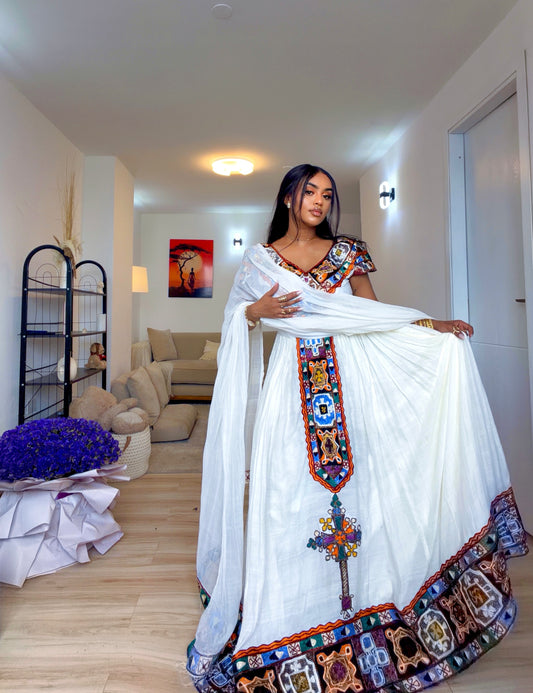 A woman models a traditional Ethiopian white georgette Ethiopian clothing with colorful embroidered details and a patterned border, standing in a modern living room.