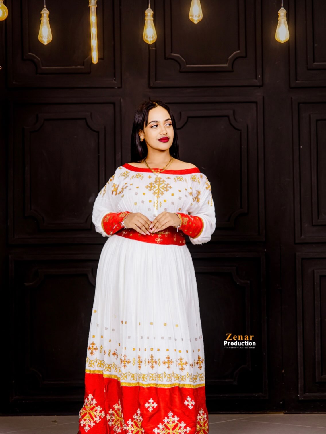 A woman modeling a traditional white, red, and gold embroidered Habesha Dress against a dark wood-paneled wall with hanging light bulbs.