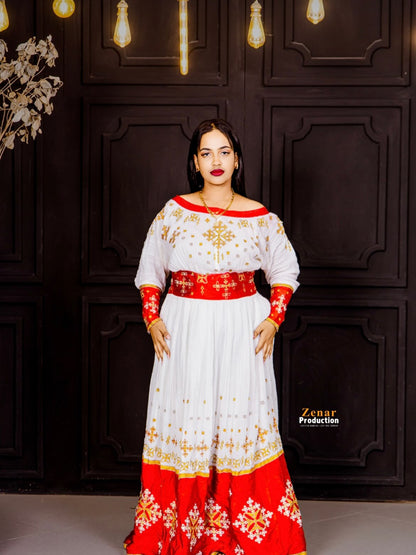 A woman modeling a traditional off-the-shoulder white Habesha Dress with red and gold embroidery, a red waist sash, and a red hem with white floral patterns, standing in front of a dark wood paneled wall.