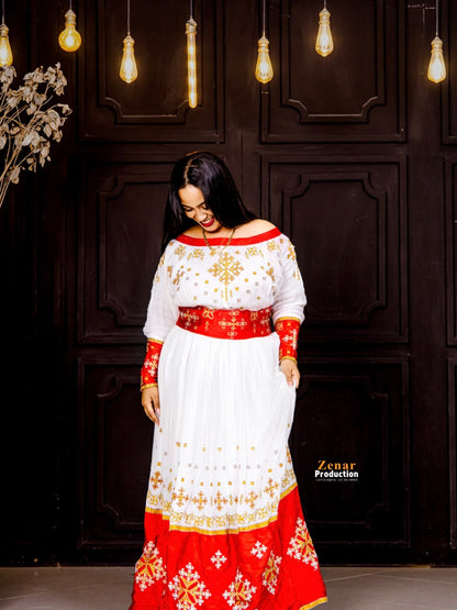 A woman wearing a traditional off-the-shoulder white and red Habesha Dress with gold embroidery, standing in front of a dark wooden paneled wall with hanging light bulbs.