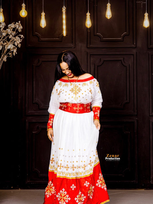 A woman wearing a traditional off-the-shoulder white and red Habesha Dress with gold embroidery, standing in front of a dark wooden paneled wall with hanging light bulbs.