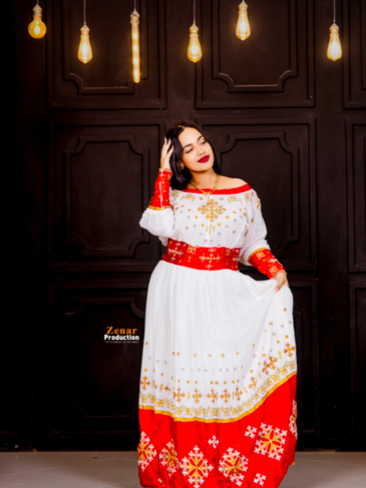 A woman models a traditional white, red, and gold embroidered Habesha Dress against a dark wood-paneled wall with hanging light bulbs.