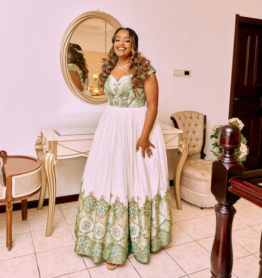 A woman wearing a formal floor-length Habesha Dress stands indoors in a decorated room with a console table, mirror, chairs, and tiled floor. The dress has a white skirt with ornate green and teal floral embroidery concentrated on the bodice and lower hem.